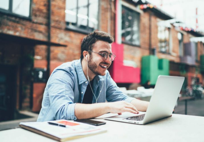 Satisfied young male in trendy casual wear and eyeglasses having fun while surfing on laptop with earphones at outdoors terrace of cafe